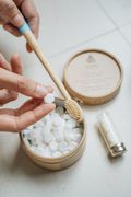 A hand tipping a toothpaste tablet from a Dentos tin beside a bamboo toothbrush