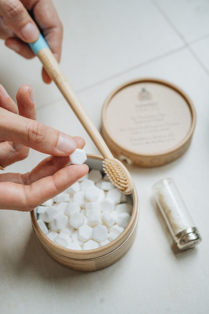 A hand tipping a toothpaste tablet from a Dentos tin beside a bamboo toothbrush