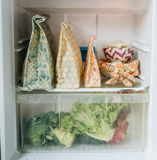 Three SuperBee beeswax bags standing on a fridge shelf beside bowls sealed with beeswax wraps, fresh vegetables in the drawer below