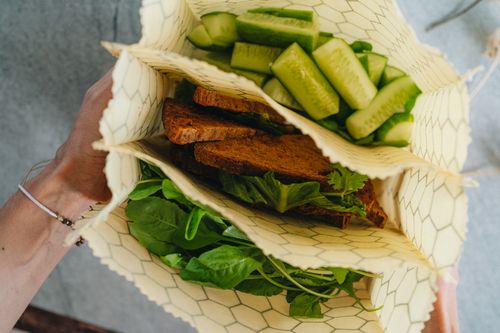 Three honeycomb-print SuperBee beeswax bags held open, packed with cucumber sticks, rye bread and salad leaves