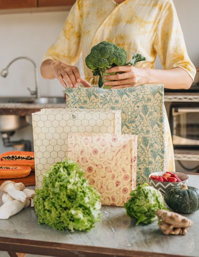 A woman packing broccoli into a SuperBee beeswax bag in a kitchen, with more bags and fresh vegetables on the counter