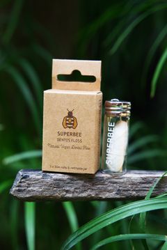A SuperBee biodegradable dental floss box and glass refill vial outdoors among green leaves