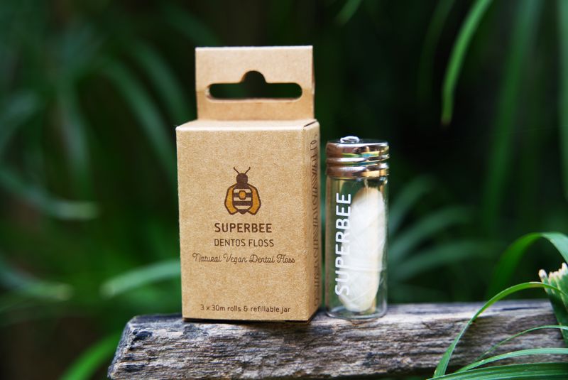 A SuperBee biodegradable dental floss box and glass refill vial outdoors among green leaves