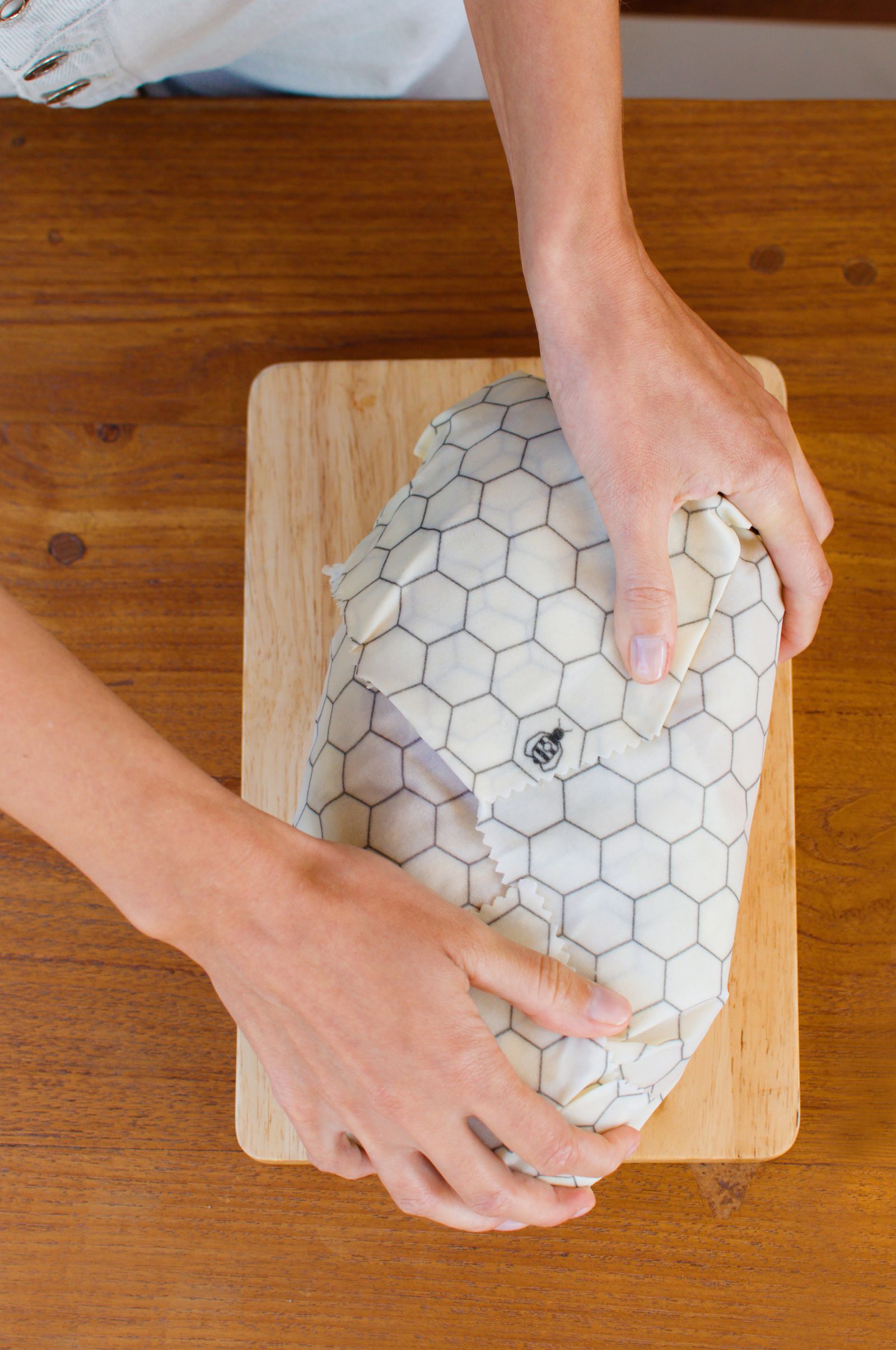 Hands folding a honeycomb-print beeswax wrap around a loaf on a board
