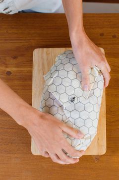Hands folding a honeycomb-print beeswax wrap around a loaf on a board