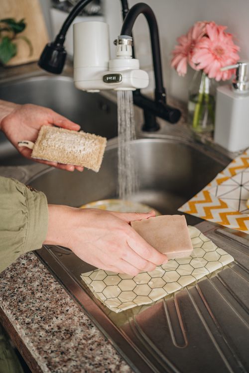 A hand lifting the dishwashing bar from a honeycomb beeswax wrap by the sink, scrubber in the other hand
