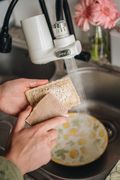 Hands rinsing the loofah scrubber and the dishwashing soap bar under a running tap