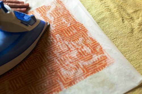 An iron pressing melted beeswax into orange-printed fabric through baking paper