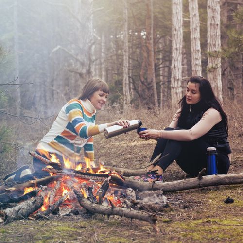 Two friends sitting by a campfire in a pine forest, sharing a flask